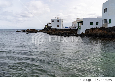 The serene coastal scene of Punta Mujeres, Lanzarote, features white buildings contrasted against dark volcanic rocks, near the tranquil sea. The serene coastal scene of Punta Mujeres, Lanzarote, features white buildings contrasted against dark volcanic rocks, near the tranquil sea. 137887069