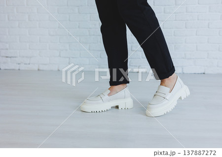 Legs of a young beautiful woman in black trousers and modern stylish white leather loafers. New collection of women's shoes spring-summer 2022 137887272