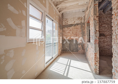 An unfinished room undergoing renovation. Exposed brick walls and drywall are visible. Natural light enters through a large window, illuminating the space. An unfinished room undergoing renovation. Exposed brick walls and drywall are visible. Natural light enters through a large window, illuminating the space. 137887759