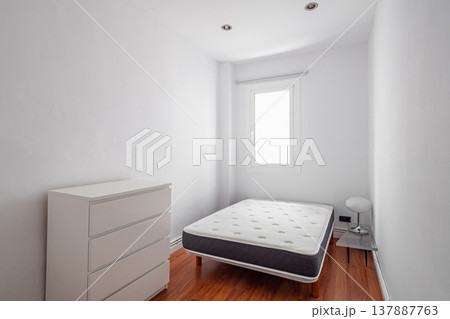 A minimalistic bedroom with a single bed and a white dresser. The room has white walls and wooden flooring, with a window allowing natural light. A minimalistic bedroom with a single bed and a white dresser. The room has white walls and wooden flooring, with a window allowing natural light. 137887763