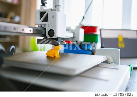 A modern 3D printer is actively creating a small yellow figurine on its build plate with filament spools in the background A modern 3D printer is actively creating a small yellow figurine on its build plate with filament spools in the background 137887825