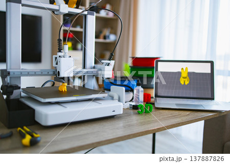 A 3D printer is actively creating a small yellow object on its build plate, with filament spools and a laptop displaying the design nearby 137887826