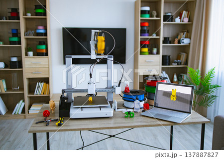 A 3D printer is actively creating a yellow object on a desk with spools of filament, tools, and a laptop displaying a 3D model 137887827