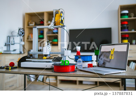 A modern 3D printer is set up on a desk with spools of filament and a laptop displaying a 3D model 137887831