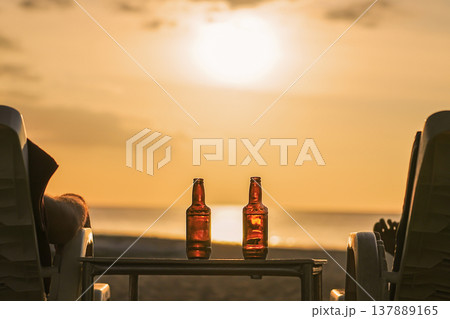 Two bottles of beer are on the table at sunset on the beach - Two friends or lovers are watching the sunset sitting in chairs back view. The concept travel and relaxing holidays with relaxing drinks Two bottles of beer are on the table at sunset on the beach - Two friends or lovers are watching the sunset sitting in chairs back view. The concept travel and relaxing holidays with relaxing drinks 137889165