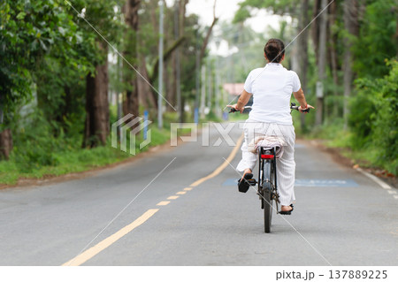 a woman riding a bicycle on an asphalt road, rear view, which passes through a wooded or park area in summer. Copy space on the left, she is dressed in light clothes. Freedom and relaxation - A quiet 137889225