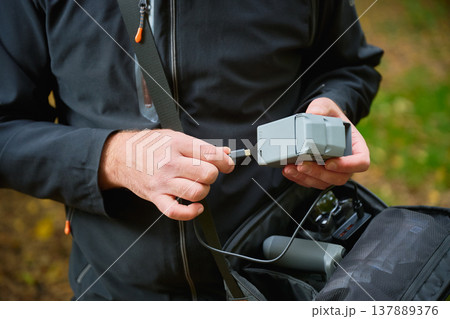 Man connects cable to drone battery for charging. Bearded man preparing power supply in park, managing equipment from bag, concept of energy, preparation and technology 137889376