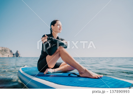 Woman Paddleboard Ocean - Young woman in wetsuit sits on paddleboard in blue water and sunny day. Woman Paddleboard Ocean - Young woman in wetsuit sits on paddleboard in blue water and sunny day. 137890993