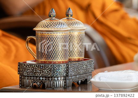 Thai ceramic cup with detailed gold and blue pattern placed on a decorative tray with a warm indoor background highlighting handcrafted design. 137892422