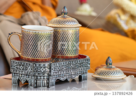Thai ceramic cup with detailed gold and blue pattern placed on a decorative tray with a warm indoor background highlighting handcrafted design. 137892426