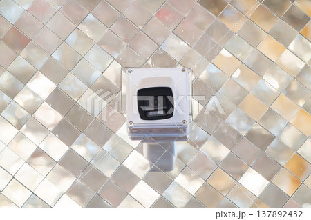 Security camera mounted on wall with reflective mosaic background. Device positioned for monitoring and surveillance in indoor space. 137892432