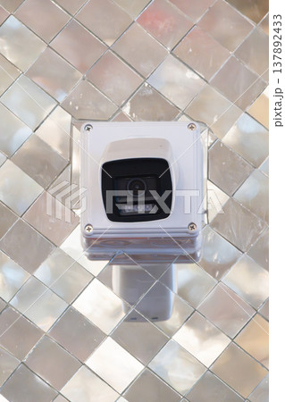 Security camera mounted on wall with reflective mosaic background. Device positioned for monitoring and surveillance in indoor space. 137892433