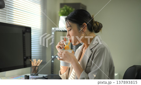 Businesswoman eating instant noodles at office desk, casual lunch break during workday in modern workspace. 137893815