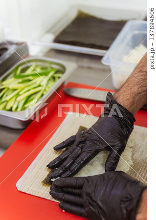 Chef Hands Preparing Nori With Cucumber On Bamboo Mat And Red Cutting Board, Thin Green Strips Chef Hands Preparing Nori With Cucumber On Bamboo Mat And Red Cutting Board, Thin Green Strips 137894606