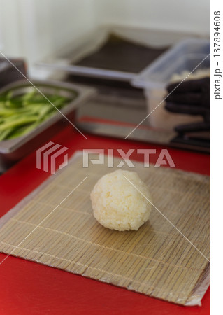 Single Rice Ball On Bamboo Mat, Resting On Red Cutting Board, Ingredient Trays Blurred In Background 137894608