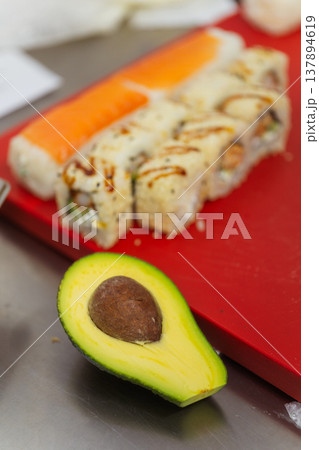 Sushi Rolls Halved Avocado On Red Board, Chef Assembly Scene With Torched Topping, Rice Grains Sushi Rolls Halved Avocado On Red Board, Chef Assembly Scene With Torched Topping, Rice Grains 137894619