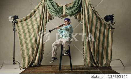 Young boy playing clarinet sitting on stool in studio stage with vintage curtains. 137895142