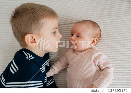 Older brother lying next to newborn baby. Tender sibling bonding moment showing curiosity, love and first interaction between children. Older brother lying next to newborn baby. Tender sibling bonding moment showing curiosity, love and first interaction between children. 137895172