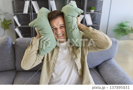 Annoyed man with ear sensitivity holding pillows to block sound of noisy neighbors 137895831