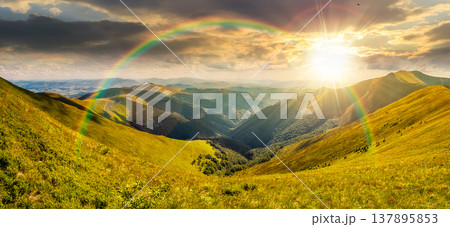 sunset over panoramic image of rolling alpine mountain ridge. gorgeous summer landscape with hillside meadows under cloudy sky. remote vista with distant valley and forested slopes under rainbow 137895853