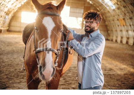 Secures the saddle. Man is with horse in the hangar Secures the saddle. Man is with horse in the hangar 137897203