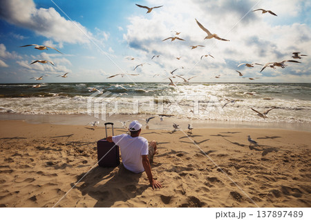 A human sits on the beach in summer with a travel suitcase. The man looks at the sea. Seagulls fly over the beach. Travel, happy holiday, summertime concept A human sits on the beach in summer with a travel suitcase. The man looks at the sea. Seagulls fly over the beach. Travel, happy holiday, summertime concept 137897489