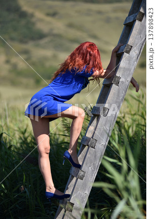 Red haired woman in vibrant blue dress and high heels ascends a rustic wooden ladder in the outdoors Red haired woman in vibrant blue dress and high heels ascends a rustic wooden ladder in the outdoors 137898423