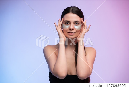 Young woman applying cosmetic eye patches for under-eye skincare, experiencing a beauty hydration treatment for wellness and self-care on a studio background 137898605