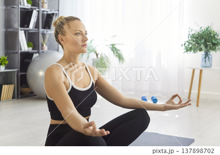 Yogi beautiful woman sitting in lotus pose on mat in home gym, meditating calmly forming mudra Yogi beautiful woman sitting in lotus pose on mat in home gym, meditating calmly forming mudra 137898702