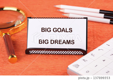 A crisp white card displaying BIG GOALS BIG DREAMS rests on a vivid orange desk, surrounded by a magnifying glass, pencils, and a calculator 137899353