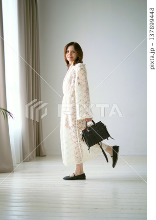 woman dressed in a white lace dress holds a black handbag while standing in a bright woman dressed in a white lace dress holds a black handbag while standing in a bright 137899448