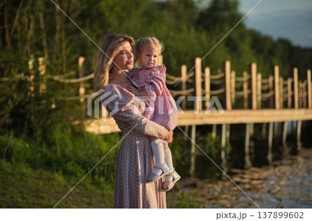 Contemplative mother holds child near lake at dusk. A tender moment evoking thoughts of protection and future dreams. Contemplative mother holds child near lake at dusk. A tender moment evoking thoughts of protection and future dreams. 137899602