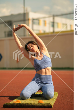 woman in blue sportswear performs a side stretch on a mat under the sunny sky. This exercise emphasizes the importance of flexibility and relaxation in a balanced fitness routine. woman in blue sportswear performs a side stretch on a mat under the sunny sky. This exercise emphasizes the importance of flexibility and relaxation in a balanced fitness routine. 137899675