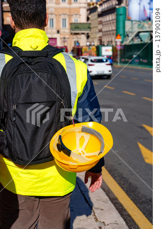 Worker walks on street with bright vest and hard hat in a busy city area during the day 137901094