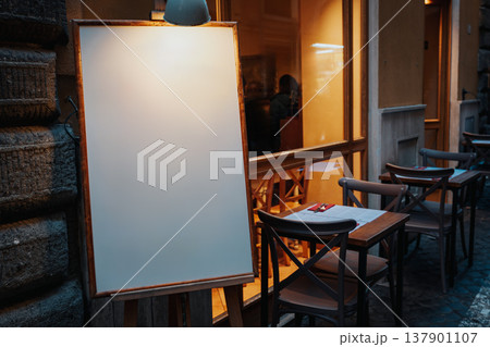 Empty board in front of cafe with tables and chairs during evening hours in city street 137901107