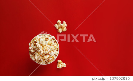 Top view of popcorn bucket and scattered kernels on red surface. Food flat lay concept. Top view of popcorn bucket and scattered kernels on red surface. Food flat lay concept. 137902050