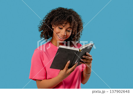 Pretty reader woman stands against bright blue backdrop, holding and reading book with curious, engaged expression. Smart girl. Discovery, learning, intellectual curiosity, spending quality time Pretty reader woman stands against bright blue backdrop, holding and reading book with curious, engaged expression. Smart girl. Discovery, learning, intellectual curiosity, spending quality time 137902648