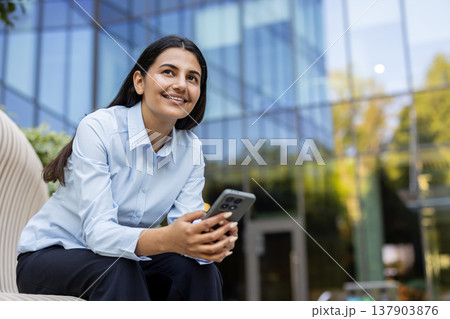 Female professional sitting on a modern bench outdoors, holding a smartphone and smiling while looking up, representing communication, innovative thinking, and career development Female professional sitting on a modern bench outdoors, holding a smartphone and smiling while looking up, representing communication, innovative thinking, and career development 137903876