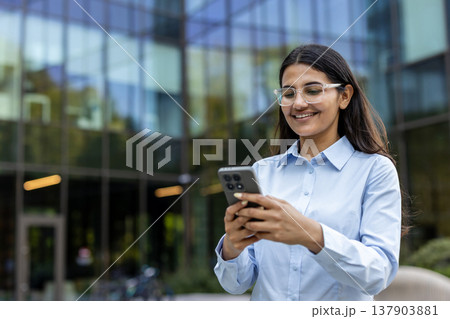 Happy young indian businesswoman reading or sending messages on her cellphone while standing outdoors in front of a modern glass office building, representing mobile communication and technology Happy young indian businesswoman reading or sending messages on her cellphone while standing outdoors in front of a modern glass office building, representing mobile communication and technology 137903881