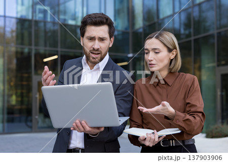 Business people standing outdoors, looking at a laptop with confused and stressed expressions, experiencing unexpected technical issues or a significant professional mistake 137903906