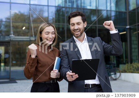 Two cheerful business professionals, a woman holding a notebook and a man holding a clipboard, celebrating their victory together with fists pumped up outside a modern office building Two cheerful business professionals, a woman holding a notebook and a man holding a clipboard, celebrating their victory together with fists pumped up outside a modern office building 137903909