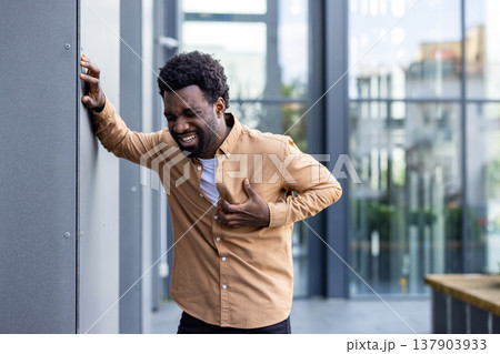 Adult individual experiencing sharp pains in their chest area, grasping their shirt while leaning against a wall outdoors, indicating discomfort and potential health suffering 137903933