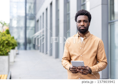 African executive standing outside a modern office building, holding a digital tablet with both hands, representing business professionalism, mobile technology, and urban connectivity African executive standing outside a modern office building, holding a digital tablet with both hands, representing business professionalism, mobile technology, and urban connectivity 137903941