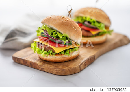 Two Beef burgers with tomato, cheese, and lettuce on a wooden board on a white table Two Beef burgers with tomato, cheese, and lettuce on a wooden board on a white table 137903982