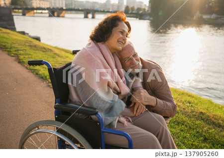 Senior woman in wheelchair walking with caregiver old man on road in park. Elderly family couple man supporting embracing woman in chair for people with disability outdoor. Rehabilitation Senior woman in wheelchair walking with caregiver old man on road in park. Elderly family couple man supporting embracing woman in chair for people with disability outdoor. Rehabilitation 137905260