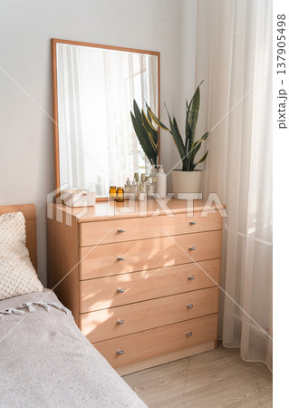 Wooden dresser in a modern bedroom interior, displaying a mirror, decorative plant, and various beauty bottles on top Wooden dresser in a modern bedroom interior, displaying a mirror, decorative plant, and various beauty bottles on top 137905498