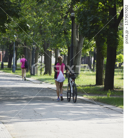 a beautiful girl walks along a path in the park and rolls a bicycle next to her 137907002