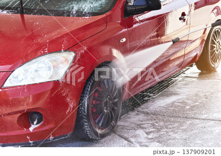 Front light of red car washed in self serve carwash, liquid reflected on side door, high pressure jet water stream spraying on wheels 137909201