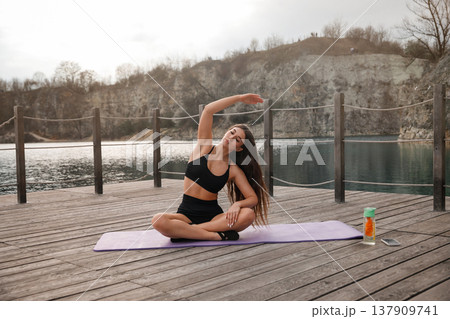 Young Woman Practicing Seated Yoga Stretch On Mat At Lake. Flexibility And Balance Training Concept Young Woman Practicing Seated Yoga Stretch On Mat At Lake. Flexibility And Balance Training Concept 137909741