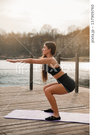 Young Fit Woman Doing Squat With Arms Extended Forward At Lake. Coordination And Strength Training Concept Young Fit Woman Doing Squat With Arms Extended Forward At Lake. Coordination And Strength Training Concept 137909760